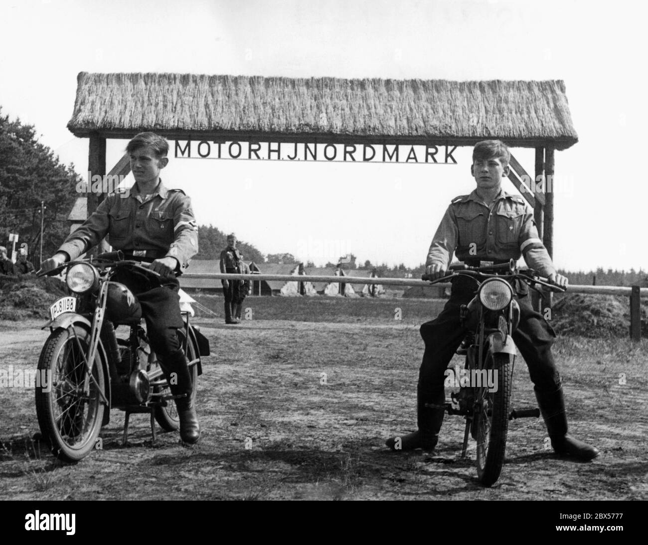 Auf dem Areal Potsdamer Platz in Breitenburg machen Mitglieder der Hitlerjugend Übungsfahrten auf ihren Motorrädern vom Typ DKW KS 200. Sie passieren einfach das Tor zum Trainingsbereich. Das Eingangstor mit der Schranke ist mit dem Namen Motor HJ Nordmark gekennzeichnet. Im Hintergrund sind Zelte. Stockfoto