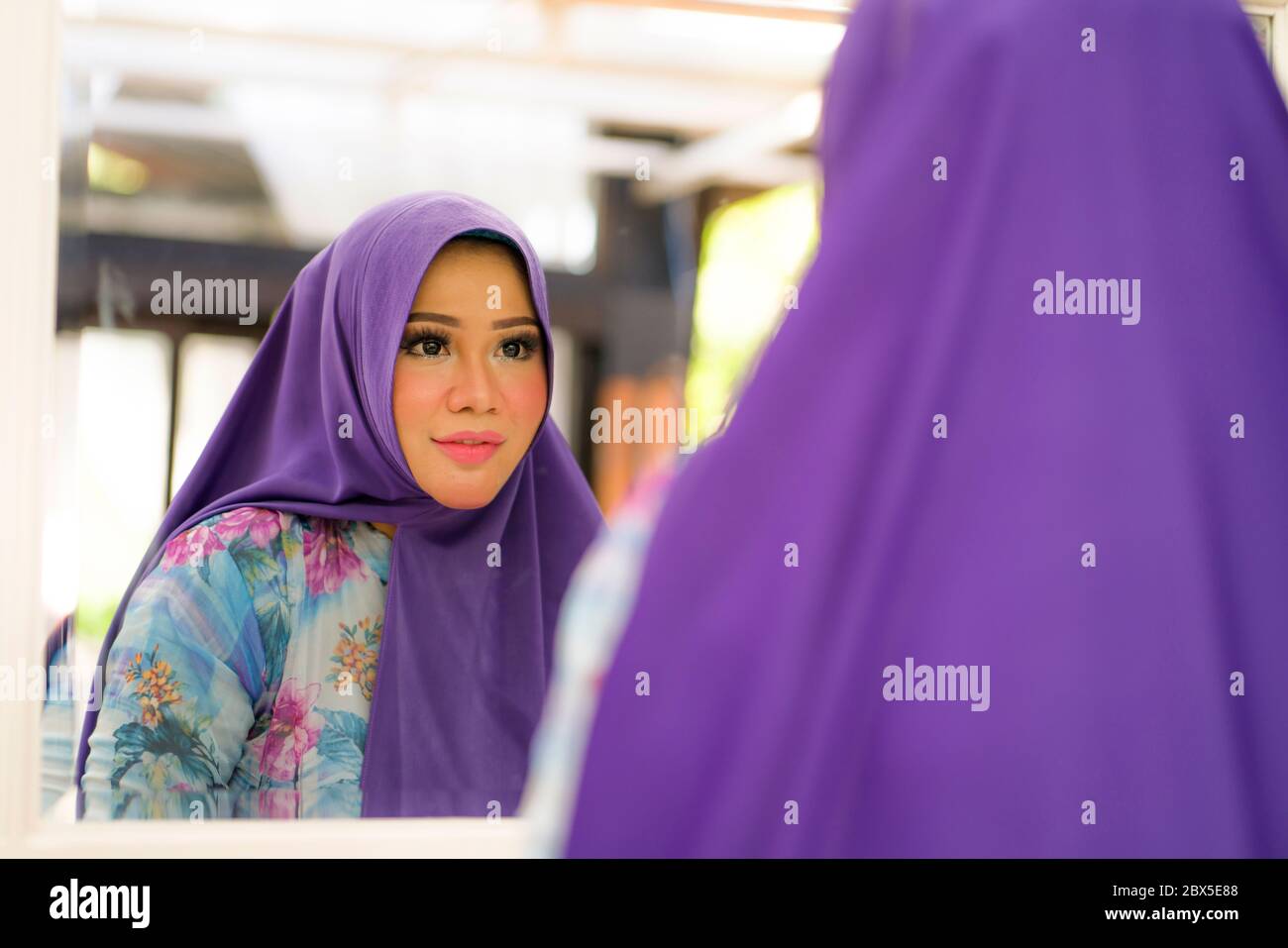 Spiegelbild der jungen glücklichen und schönen Muslimin in traditionellen Hijab Anpassung ihrer blauen Kopftuch fröhlich puppen in Schönheit und Fash Stockfoto