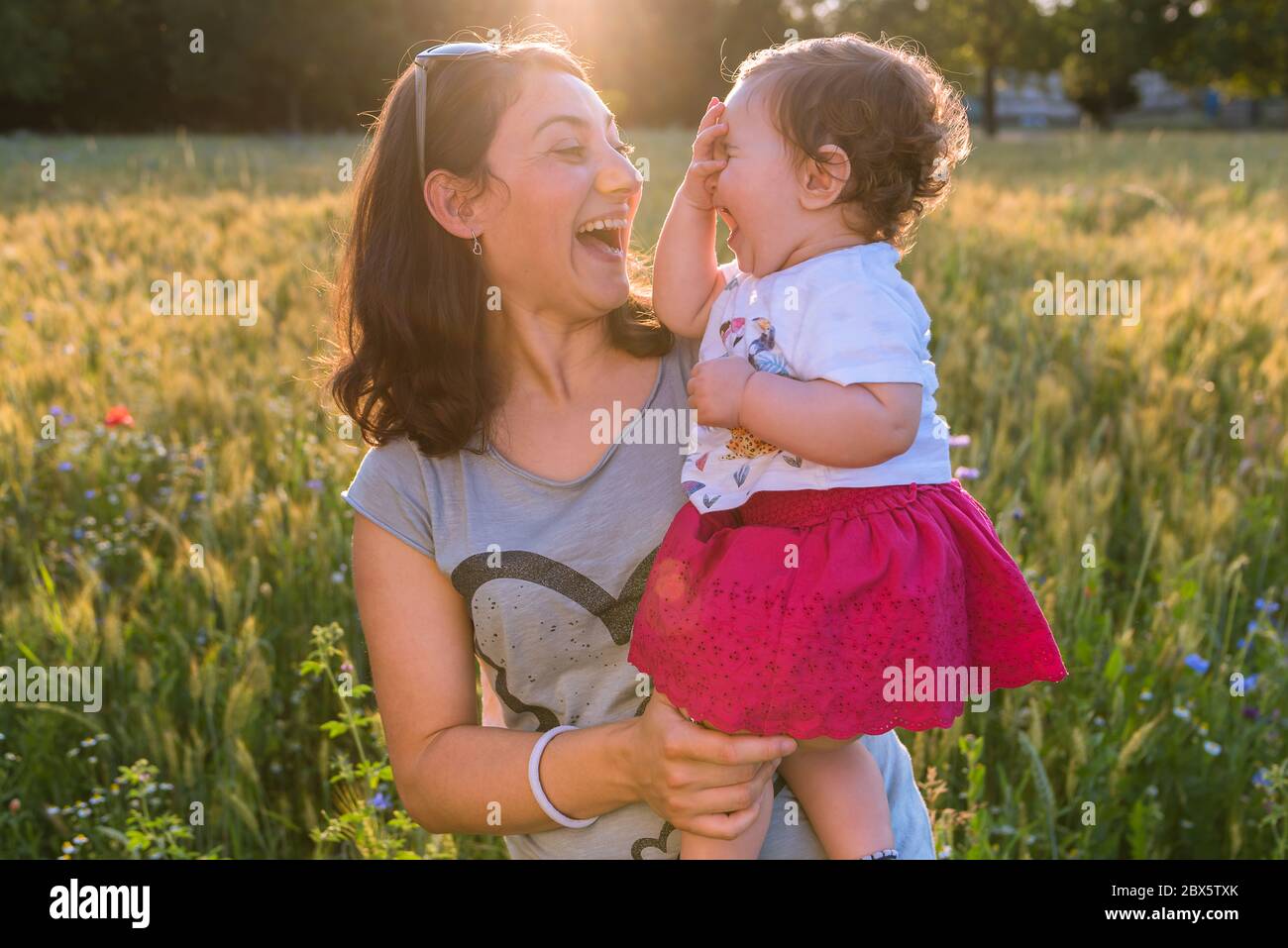 Eine junge Mutter hält ihr Baby in den Armen, während sie an einem sonnigen Tag draußen zusammen lachen. Stockfoto