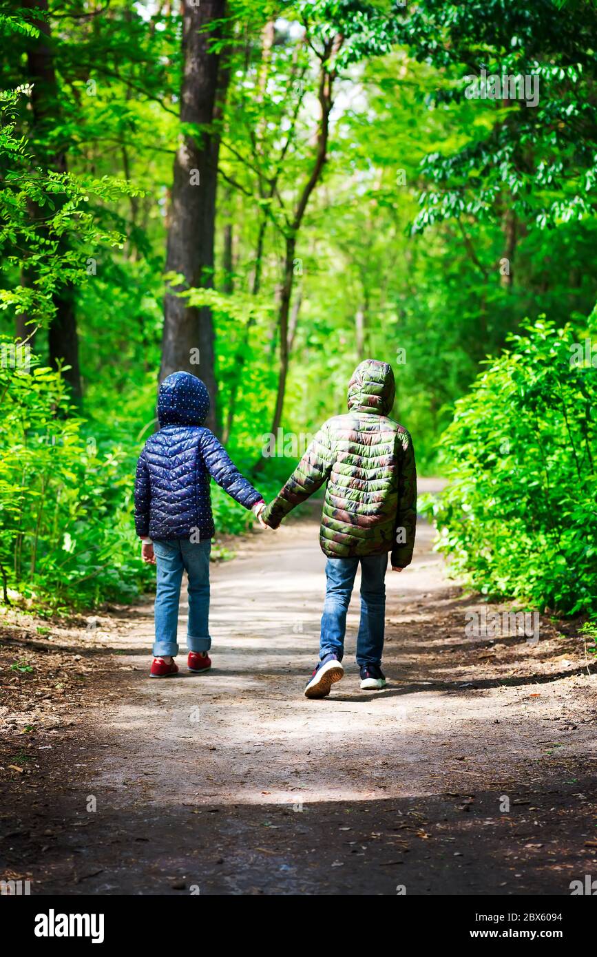 Zwei Kinder in Jacken laufen in einem sonnigen Tag in sicherer Entfernung durch den Stadtpark oder Wald und halten sich die Hände Stockfoto