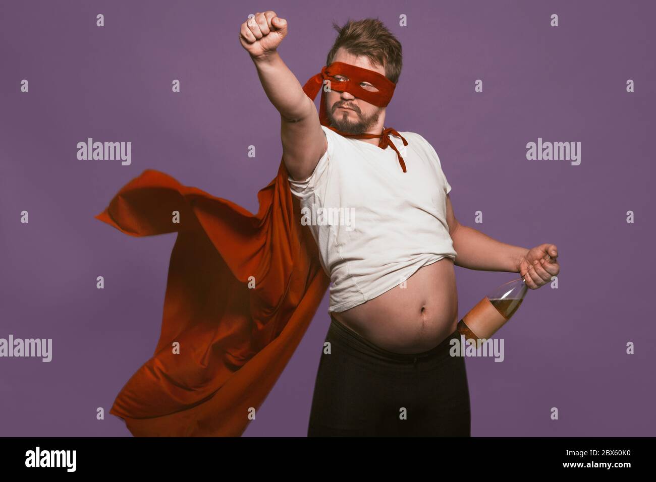 Betrunken super Antiheld hält Weinflasche zeigt seinen Bauch. Mann in einer roten Maske und einem Superheldenmantel mit Flasche Wein in einer Hand und heben seine Stockfoto