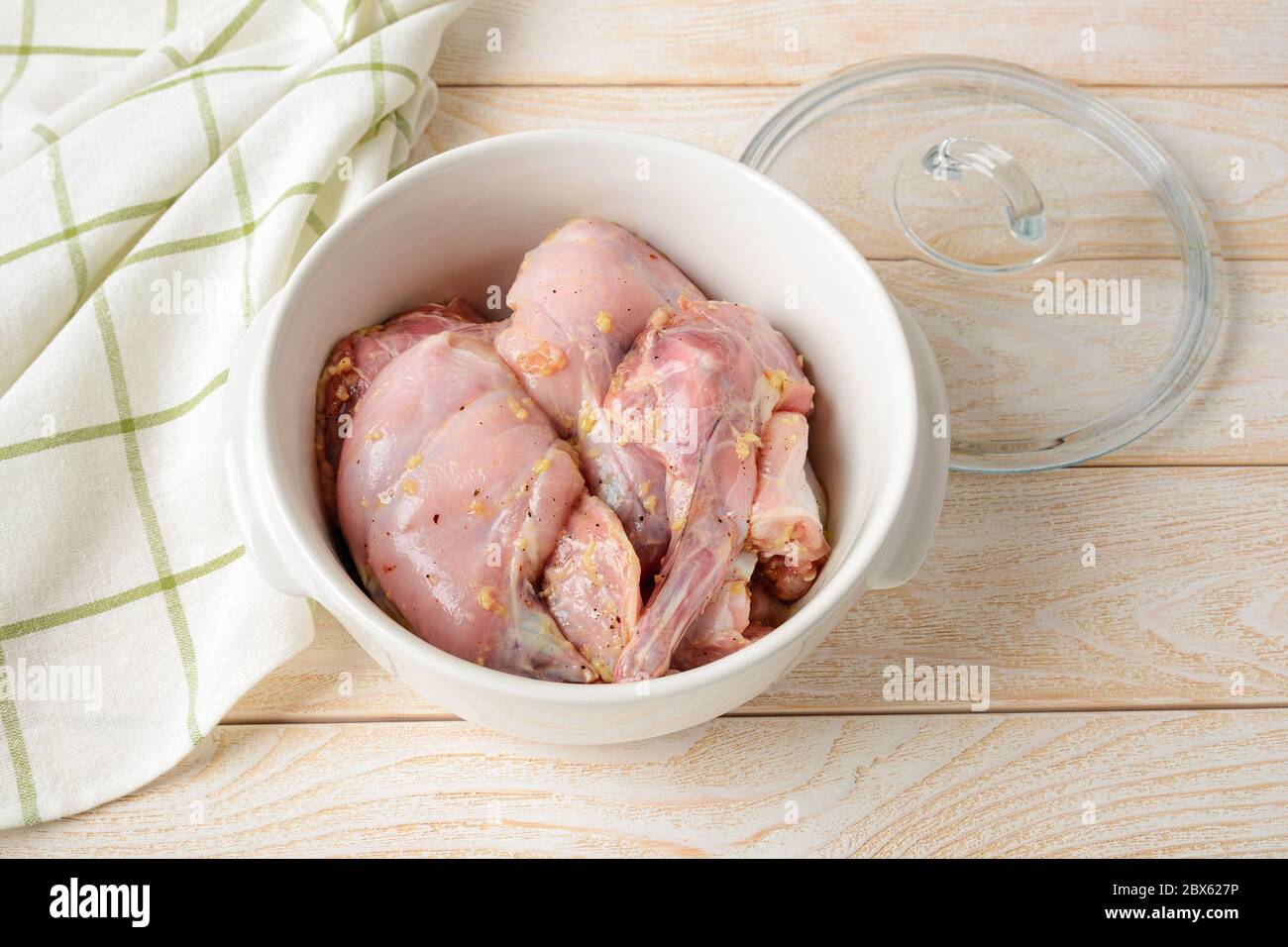Stücke von frischem rohem Kaninchenfleisch mariniert mit Olivenöl und Knoblauch vor dem Einkochen in einer weißen Keramikpfanne auf dem Küchentisch. Kochen Kaninchen Eintopf. Stockfoto