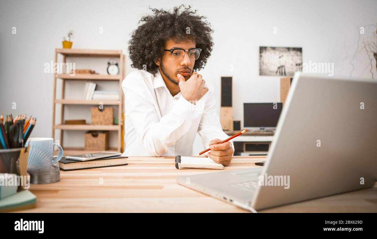Nachdenklicher junger Mann ist zu Hause vor dem Computer engagiert. Arabischer Student oder Geschäftsmann, der Notizen in einem Notizbuch schreibt. Konzept für die häuslichen Bildung. Tonbild Stockfoto