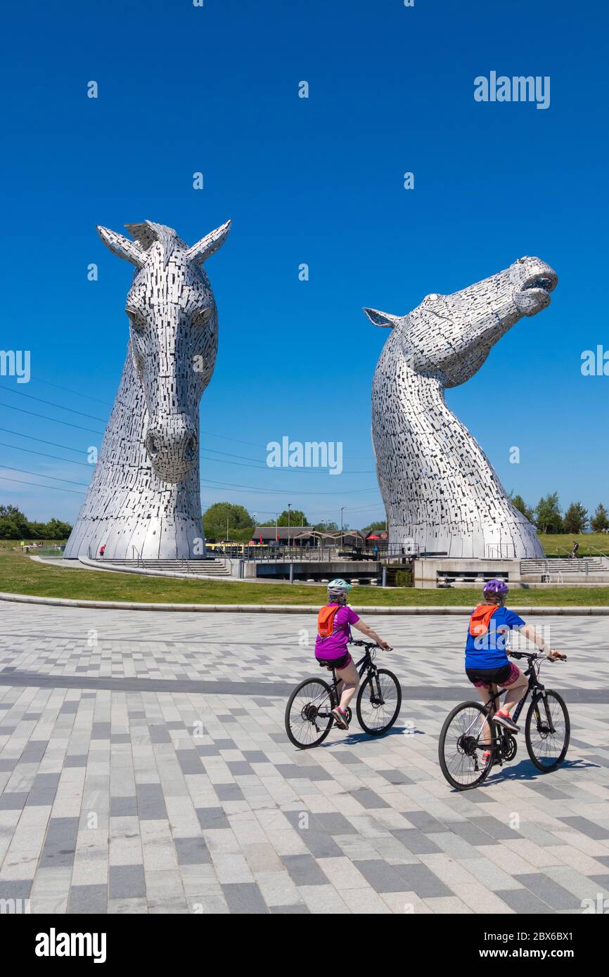 Zwei Radfahrer radeln vor den riesigen Pferdeskulpturen, die Kelpies genannt werden, in der Nähe von Falkirk, Schottland Stockfoto