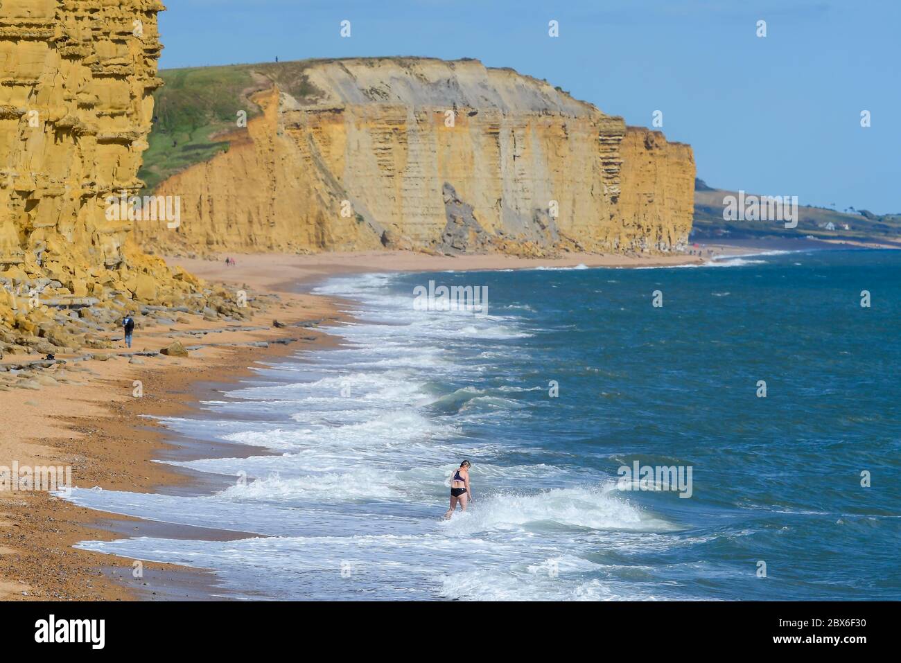 West Bay, Dorset, Großbritannien. Juni 2020. Wetter in Großbritannien. Eine Frau geht an einem sonnigen Nachmittag bei kühler Brise in das Meer am fast menschenleeren Strand unter Klippen im Seebad West Bay in Dorset. Bild: Graham Hunt/Alamy Live News Stockfoto