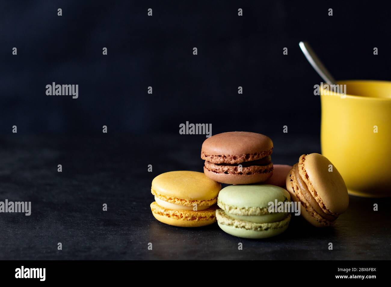 Gruppe von farbigen Macarons und gelbe Tasse auf schwarzem Hintergrund. Mit leerem Raum auf der linken Seite für Werbeplakat. Stockfoto Gruppe von farbigen Macarons und gelbe Tasse auf schwarzem Hintergrund. Mit leerem Raum auf der linken Seite für Werbeplakat. Stockfoto