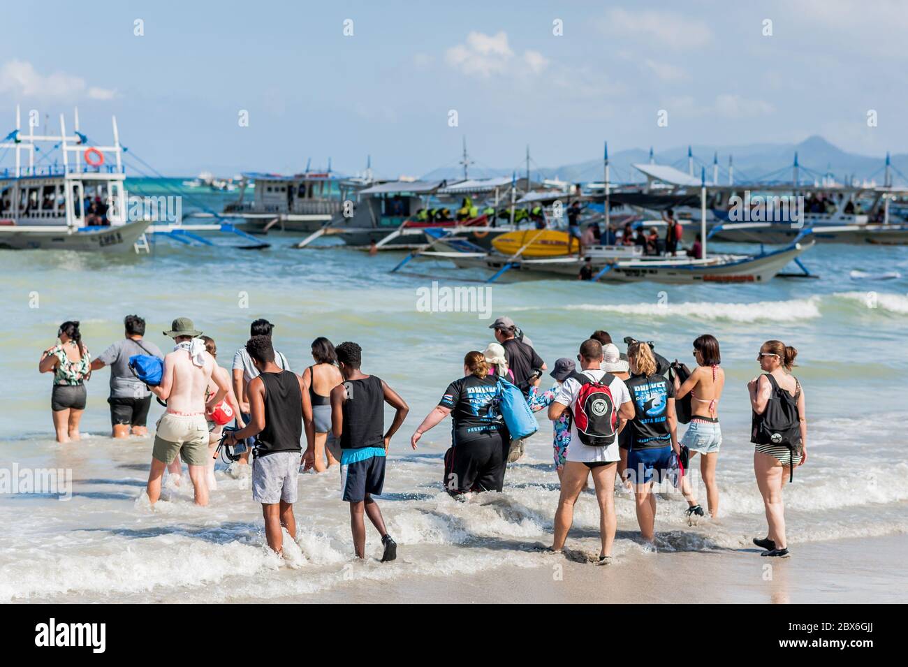 Touristen am Stadtstrand von El Nido waten zu den zahlreichen Langschwanz-Booten, die in der Brandung festgemacht sind und darauf warten, die Inseln und Strände zu besuchen. Stockfoto