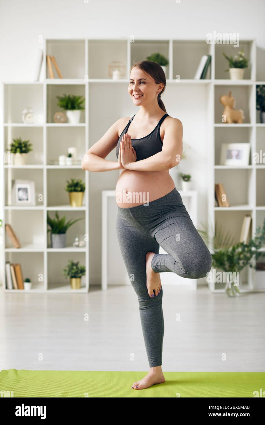 Glückliche junge schwangere Sportlerin steht auf einem Bein auf Matte mit ihren Händen zusammen von der Brust beim Yoga-Training zu Hause Stockfoto