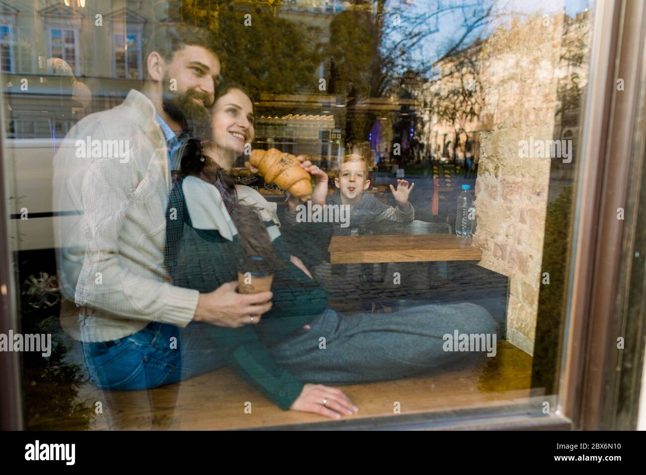 Junges Paar, das im Café sitzt und zum Fenster schaut, sich umarmt, frisch gebackene Croissants isst. Lustige kleine Junge Sohn zeigt Gesten und Stockfoto