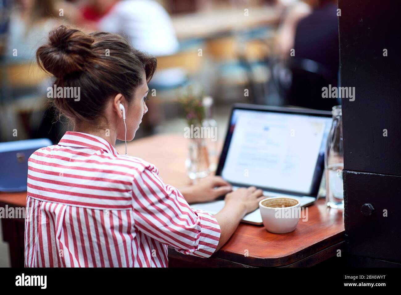 Junge Frau, die auf ihrem Laptop mit Kopfhörern in Ohren an öffentlichen Ort tippen Stockfoto