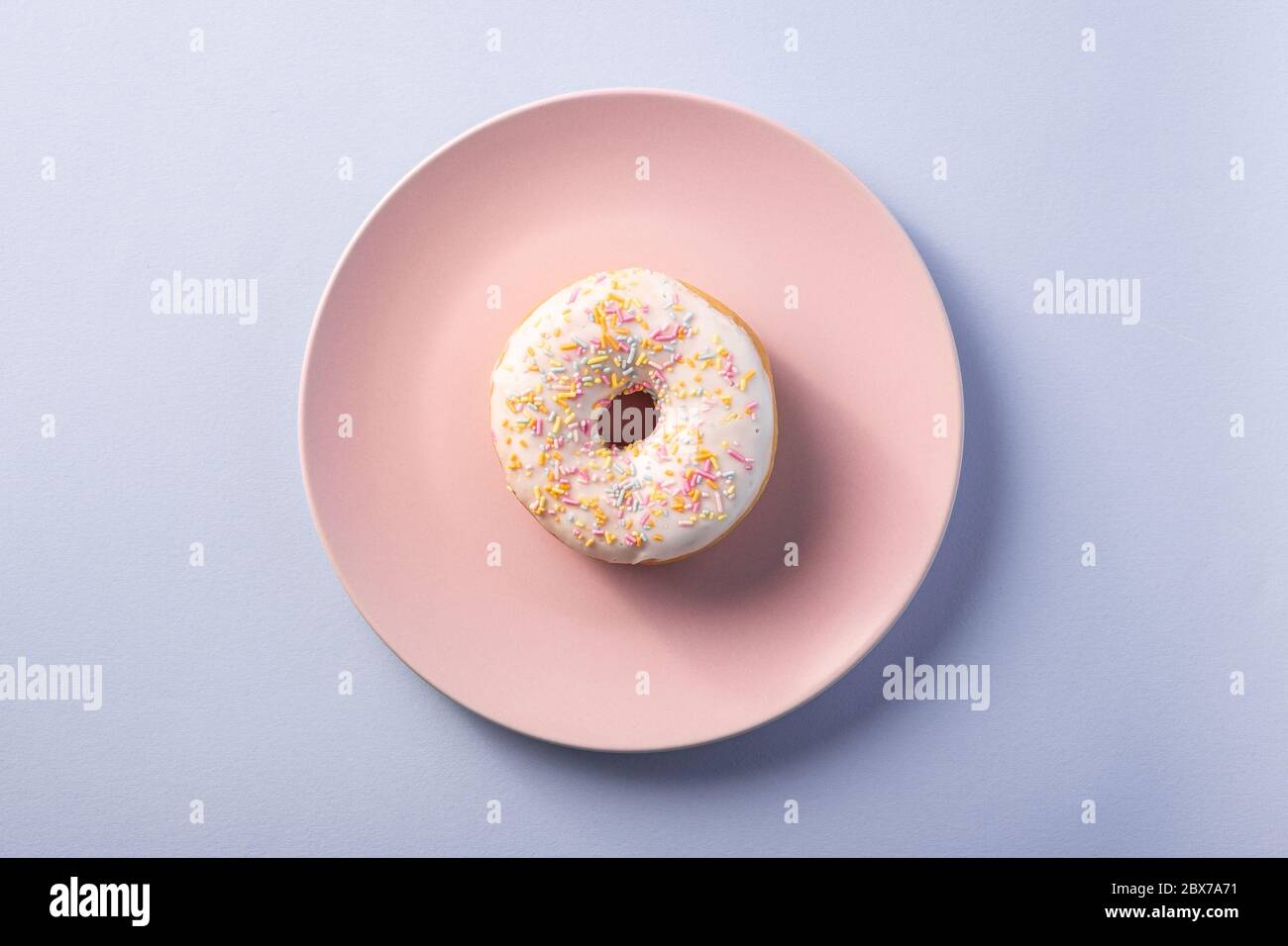 Vanilledonut mit Streuseln auf rosa Teller, süß glasierte Dessertspeise auf blauem minimalem Hintergrund, Draufsicht Stockfoto