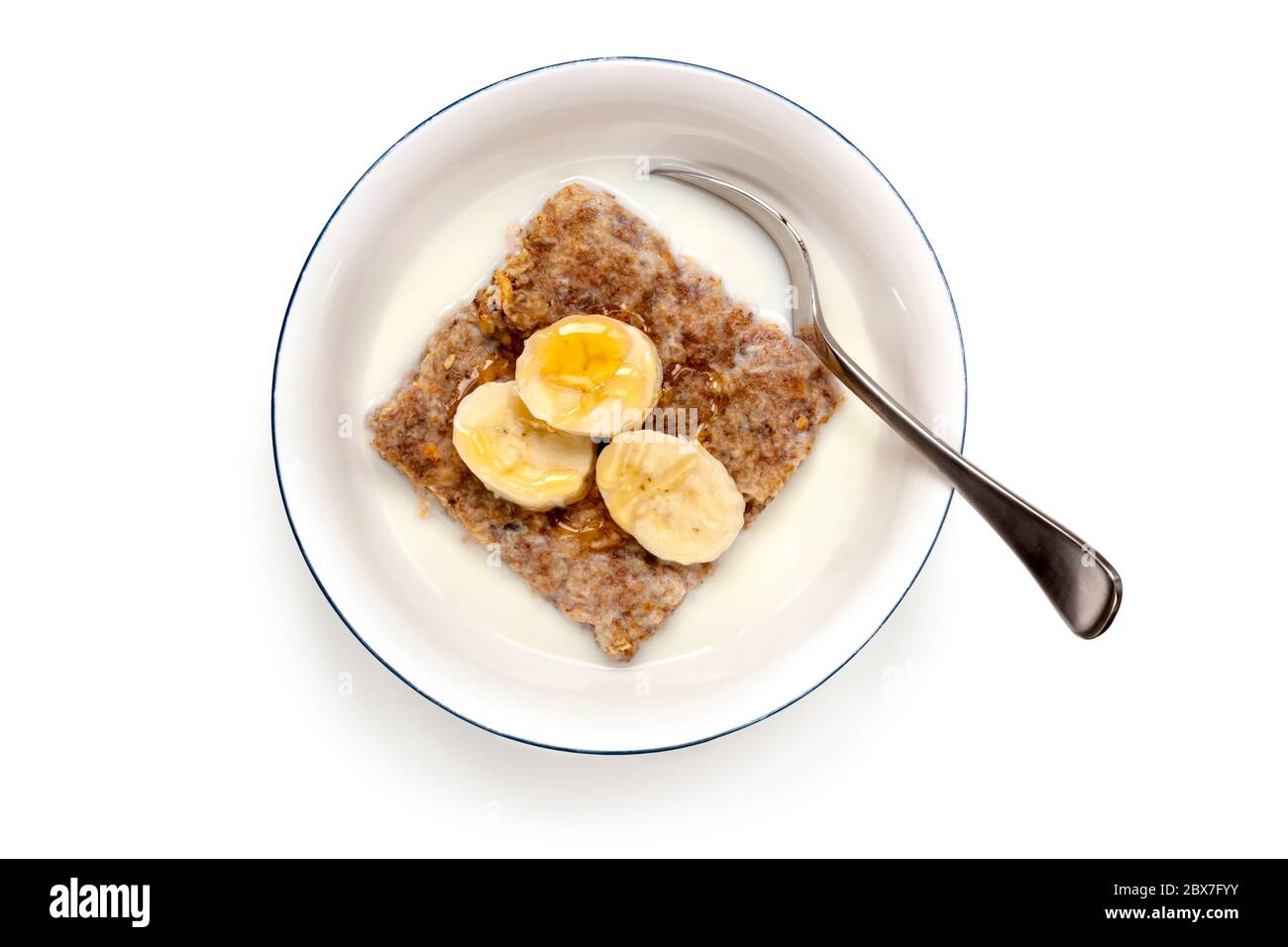 Schüssel Frühstückszerealien mit Löffel. Draufsicht, isoliert auf Weiß. Weizenkekse mit Banane und Honig gekrönt. Stockfoto