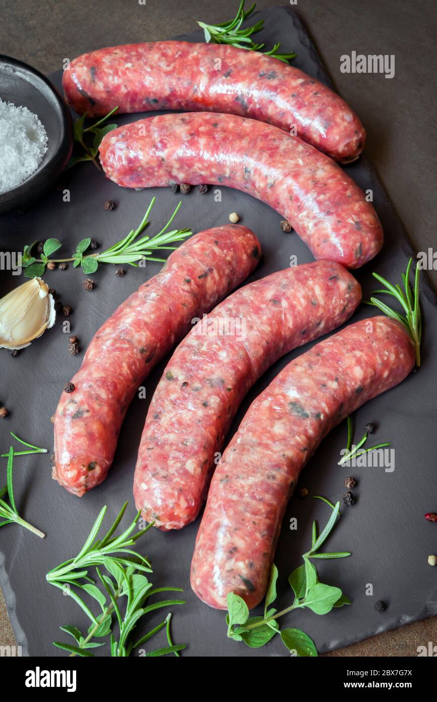 Rohe Gourmet-Würste mit frischen Kräutern und Gewürzen, auf dunklem Schiefer. Stockfoto