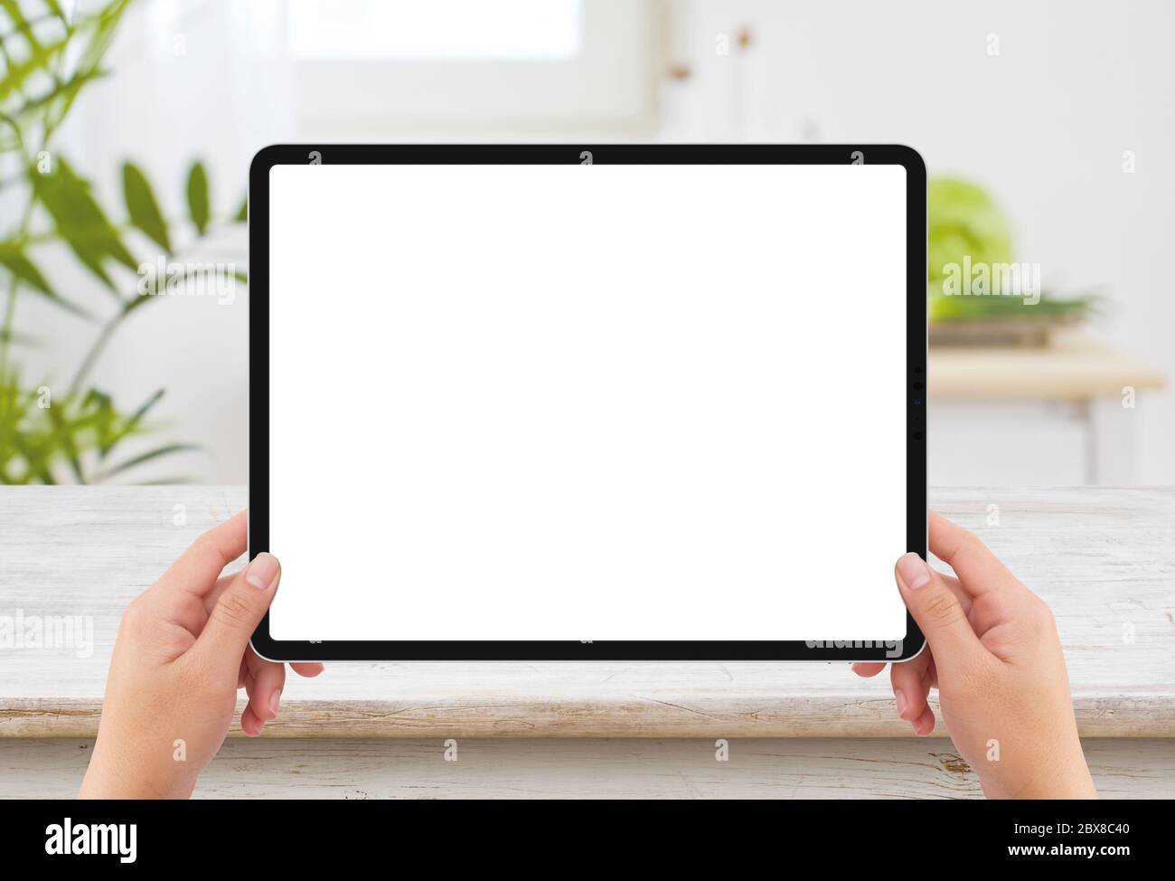 Isolierte menschliche linke Hand hält schwarze Tablet-Mediengerät mit weißen leeren Bildschirm Mockup und Holztisch in der Küche Stockfoto