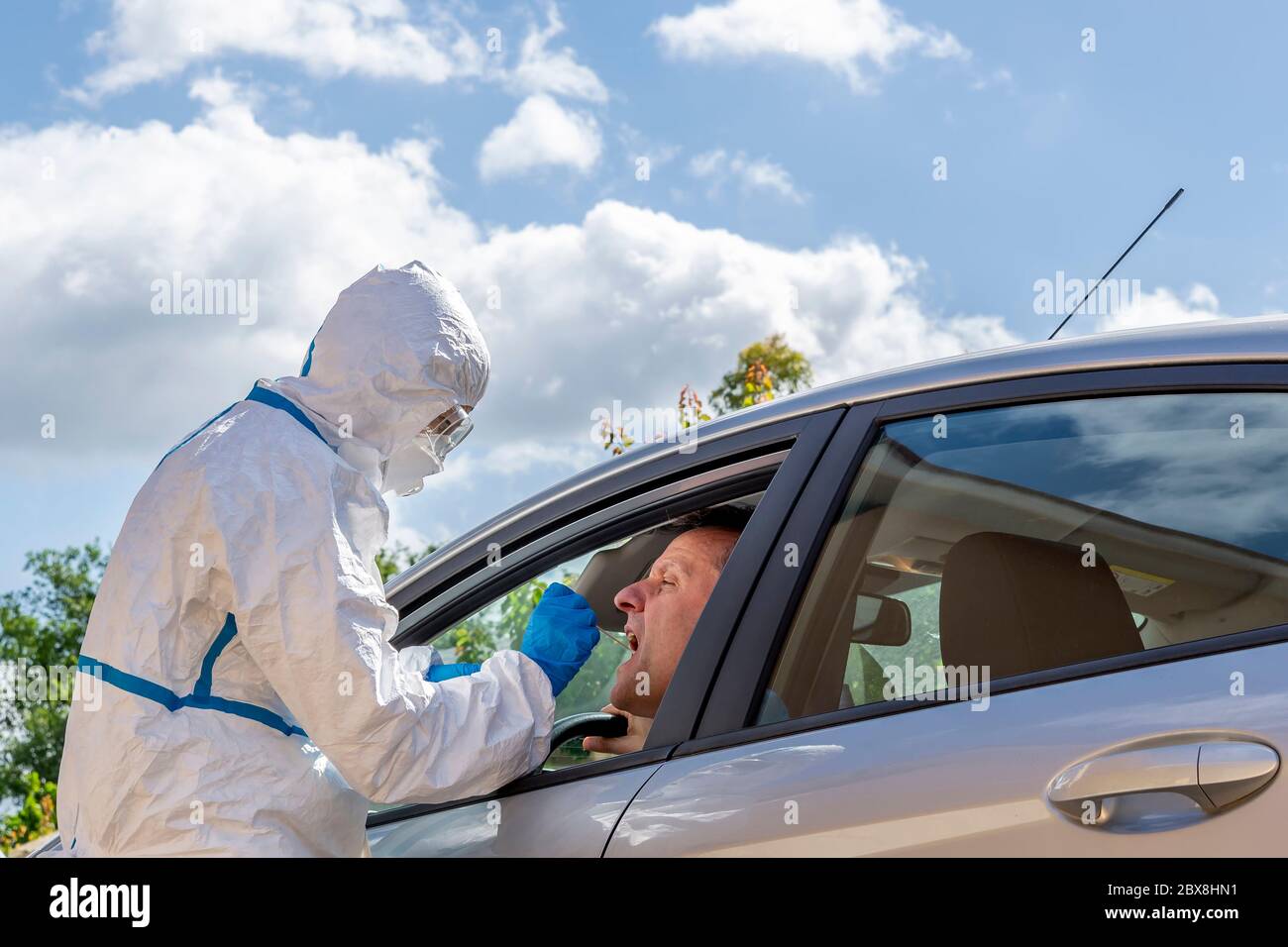 Ein Gesundheitsarbeiter, der vollständig mit einem weißen Schutzanzug und einer Brille bedeckt ist, bedient einen Kontrollabstrich für Covid-19 für einen Mann, der in seinem Auto sitzt Stockfoto