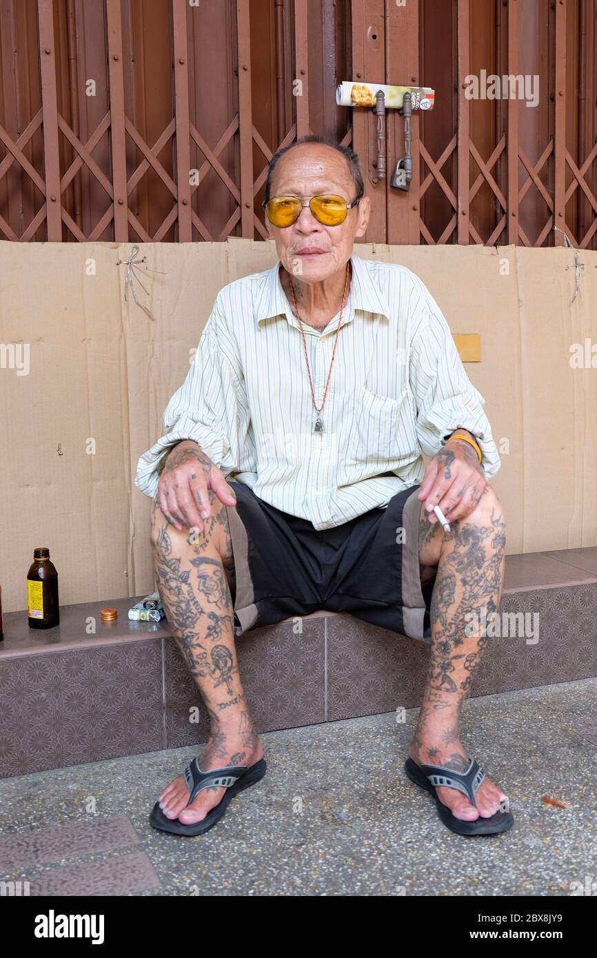 Ältere tätowierte Mann auf Schritt des geschlossenen Laden mit einem Rauch sitzen, Talat Noi, Talad Noi, Chinatown, Bangkok, Thailand, Südostasien. Stockfoto