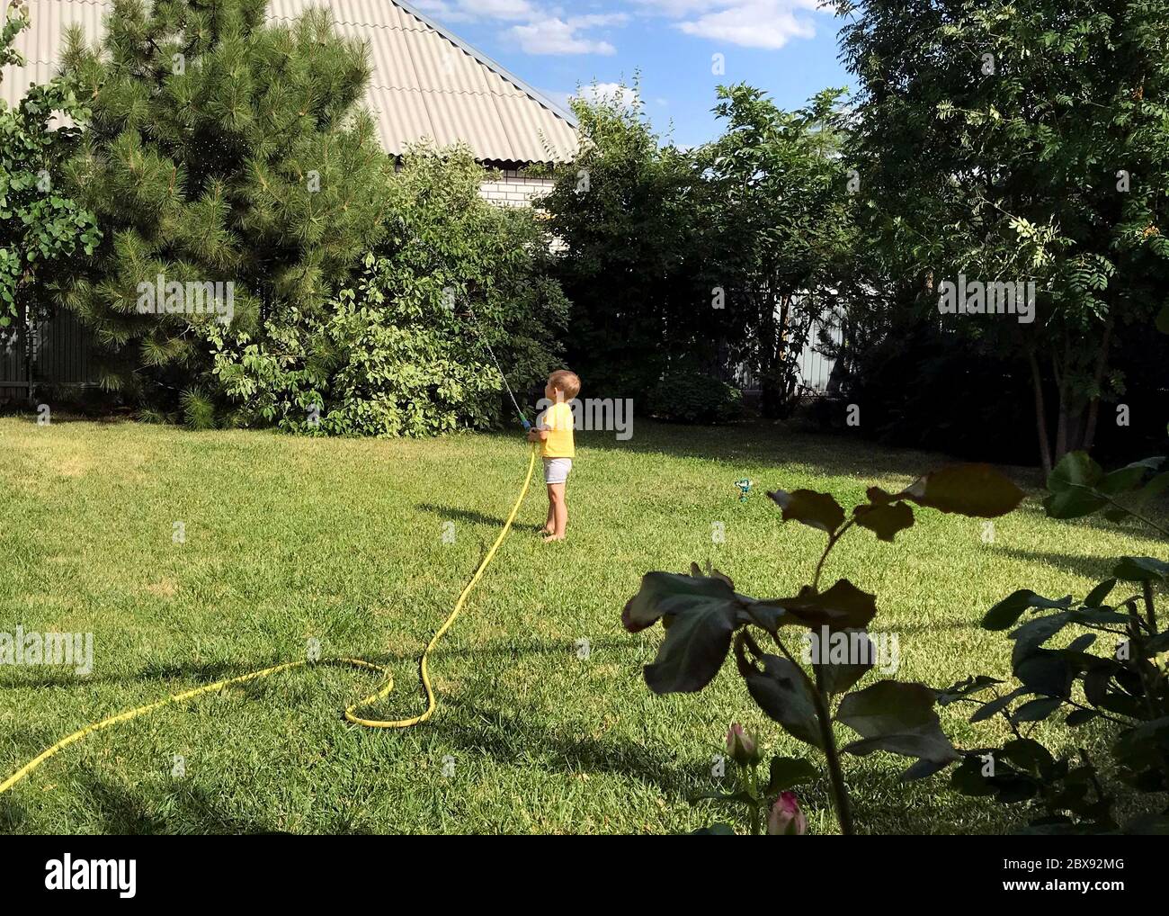 Niedliche aucasian barfeet junge zu Hause Hinterhof hält Schlauch Rohr für die Bewässerung Garten. Kind kleine Helfer spielen Gartenarbeit im Sommer im Freien Stockfoto