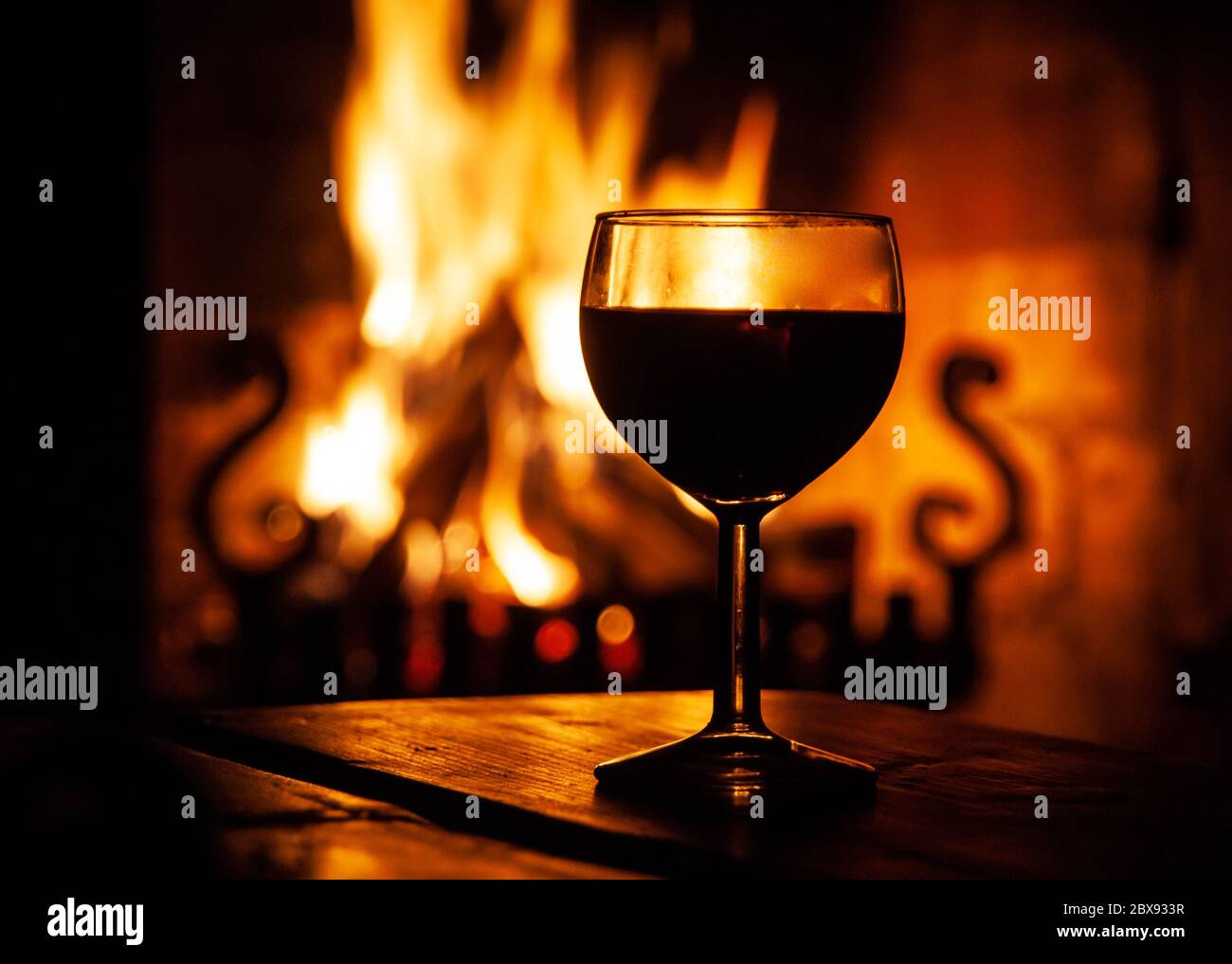 Glas Rotwein auf dem Holztisch mit brennendem Feuer im Hintergrund. Abends entspannen Sie sich an einem gemütlichen Ort. Dunkle mittelalterliche Weingüter. Stockfoto