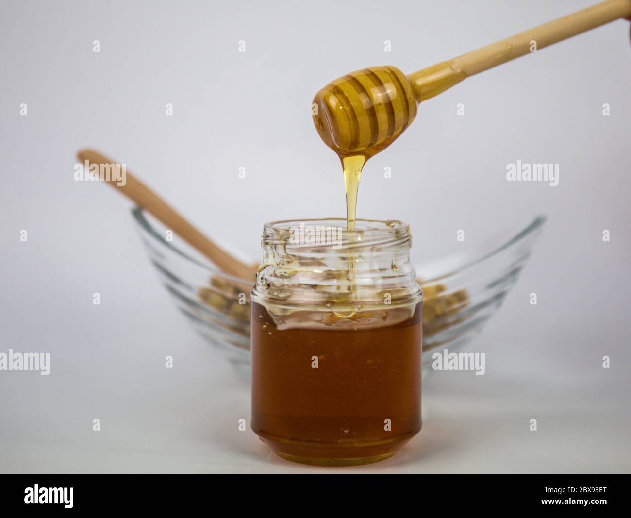 Glas mit Honig und Holzstab auf weißem Hintergrund. Aromatischen Honig in Glas gießen, Nahaufnahme. Stockfoto