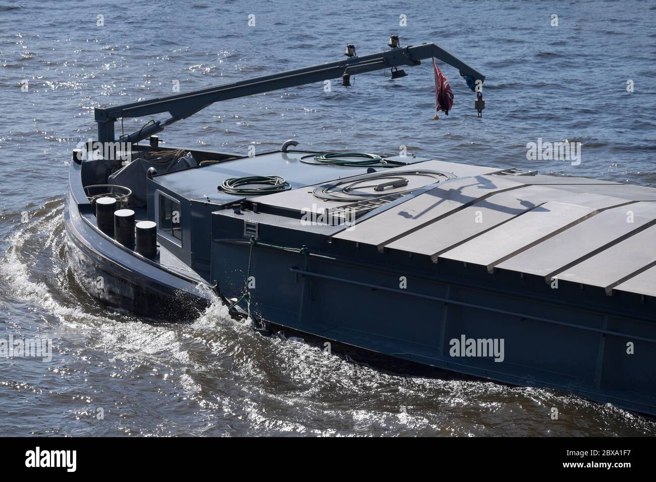 Vor einem tief liegenden Binnenschiff mit einem Kran auf dem Deck segelt durch einen Fluss. Das Wasser spritzt auf Stockfoto