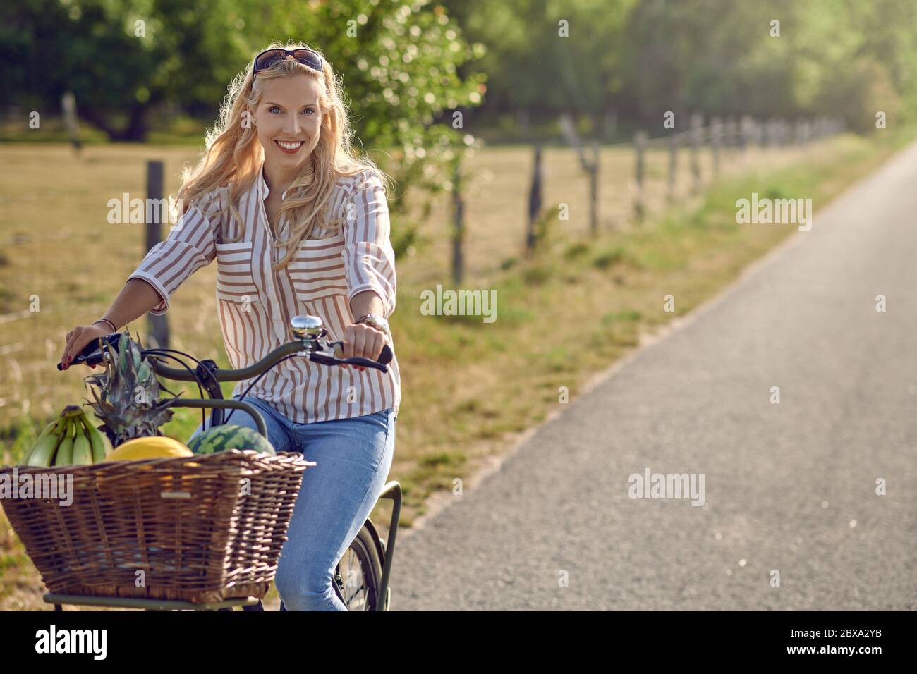 Glückliche Frau lächelnd, während Sie ein Fahrrad mit einem Korb voller frischer und gesunder Früchte in einem sonnigen Tag des Sommers auf dem Land fahren Stockfoto