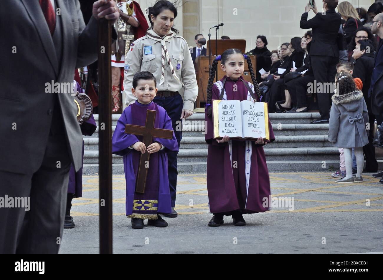 Kinder in purpurnen Kleidern stehen vor der Senglea Basilika. Der Junge auf der linken Seite hält ein Kreuz. Stockfoto