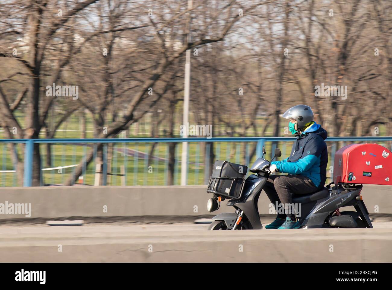 Belgrad, Serbien - 20. März 2020: Reifer Mann mit Sturzhelm, Gesichtsmaske und Handschuhen, Roller mit schwarzer Tasche im Vorderteil und roter Heckauflage Stockfoto