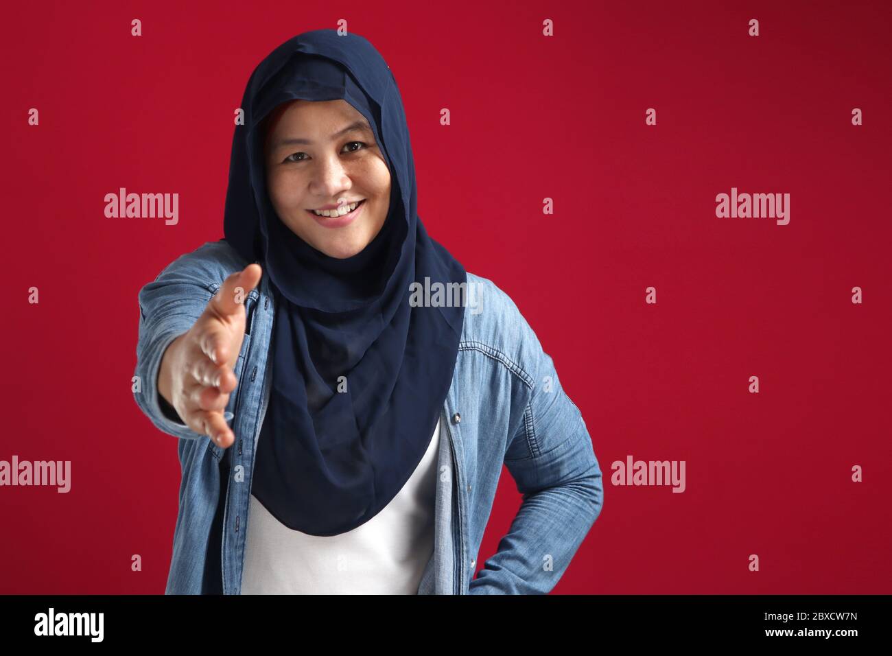 Junge schöne asiatische muslimische Dame trägt Hijab stehen über rotem Hintergrund lächelnd freundlich bietet Handschlag als Begrüßung und einladend. Erfolgreich Stockfoto