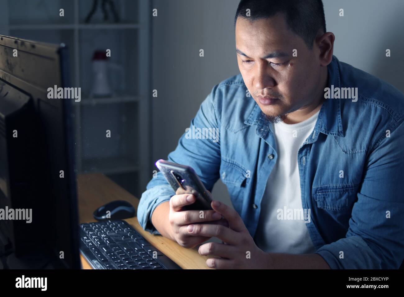 Asiatischer Mann mit Telefon-Anwendung und arbeitet auf seinem Computer in der Nacht, Online-Zahlungsbestätigung Stockfoto