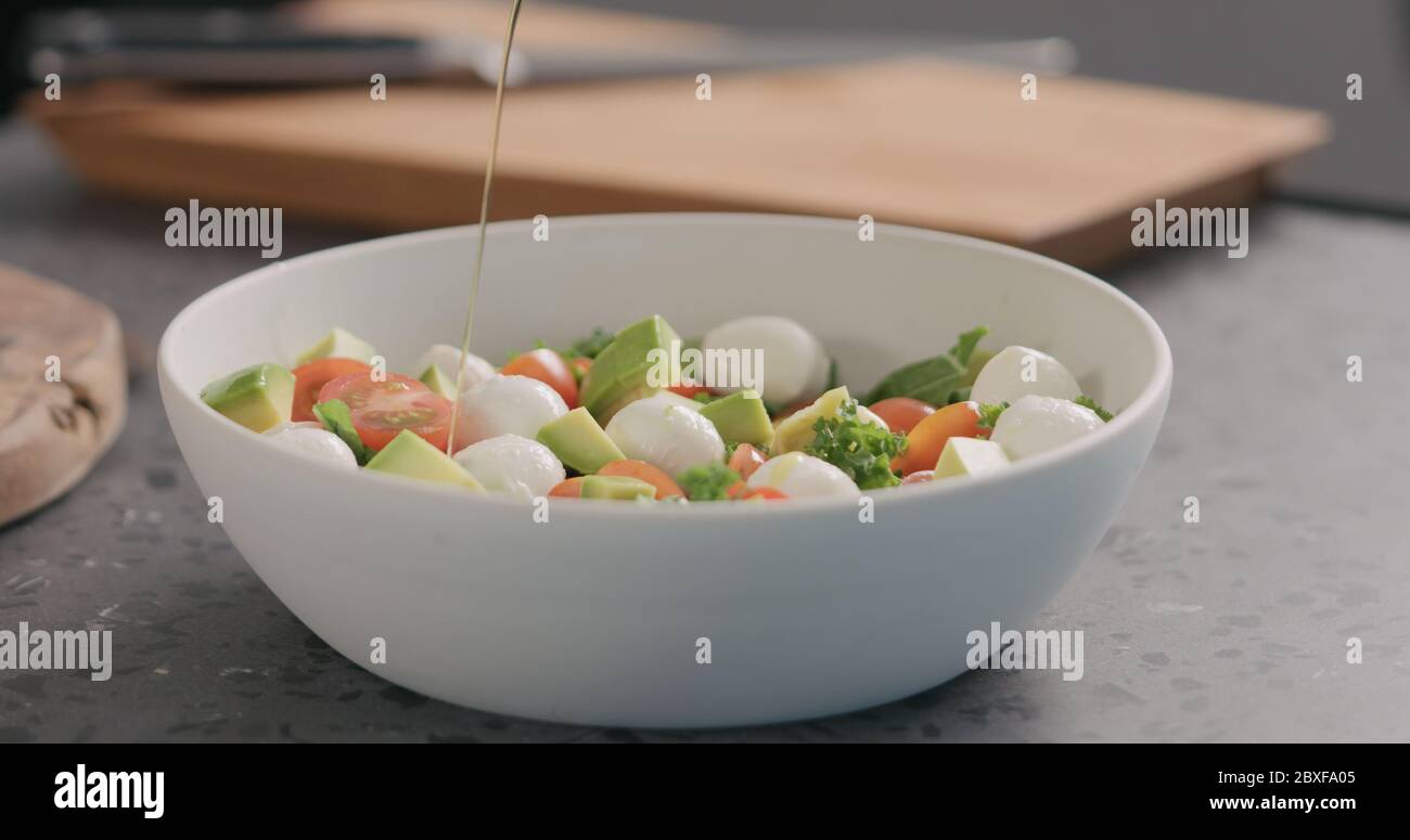 Man gibt Öl in Salat mit Grünkohl, Avocado, Mozzarella und Tomaten in einer weißen Schüssel auf der Küchenarbeitsfläche Stockfoto