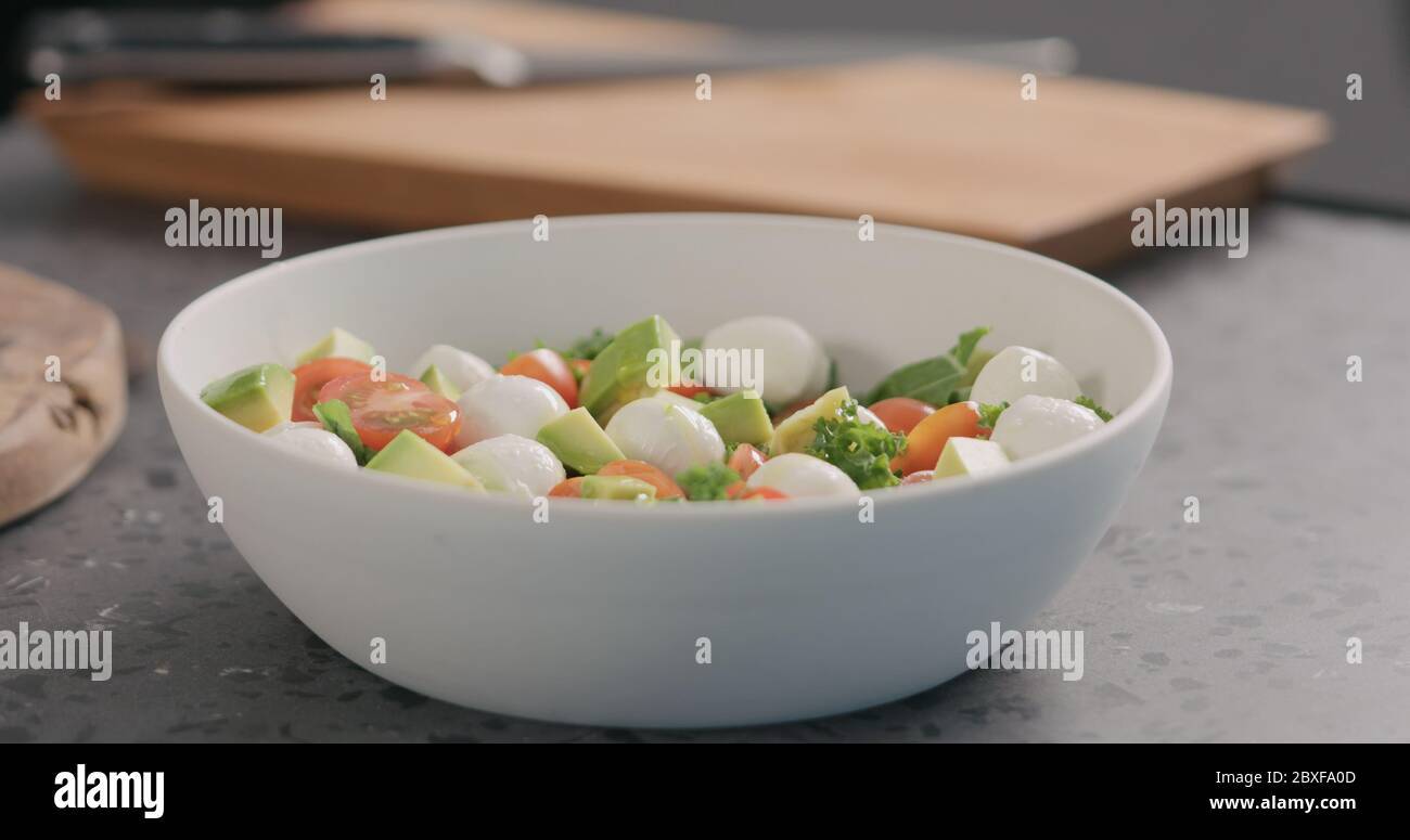 Man gibt Öl in Salat mit Grünkohl, Avocado, Mozzarella und Tomaten in einer weißen Schüssel auf der Küchenarbeitsfläche Stockfoto