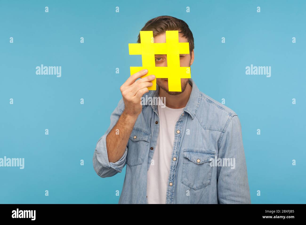 Porträt des Mannes in Denim-Shirt durch große gelbe Hashtag Symbol suchen, stehend isoliert auf blauem Hintergrund mit Kopierraum für getaggte Nachricht, Pop Stockfoto