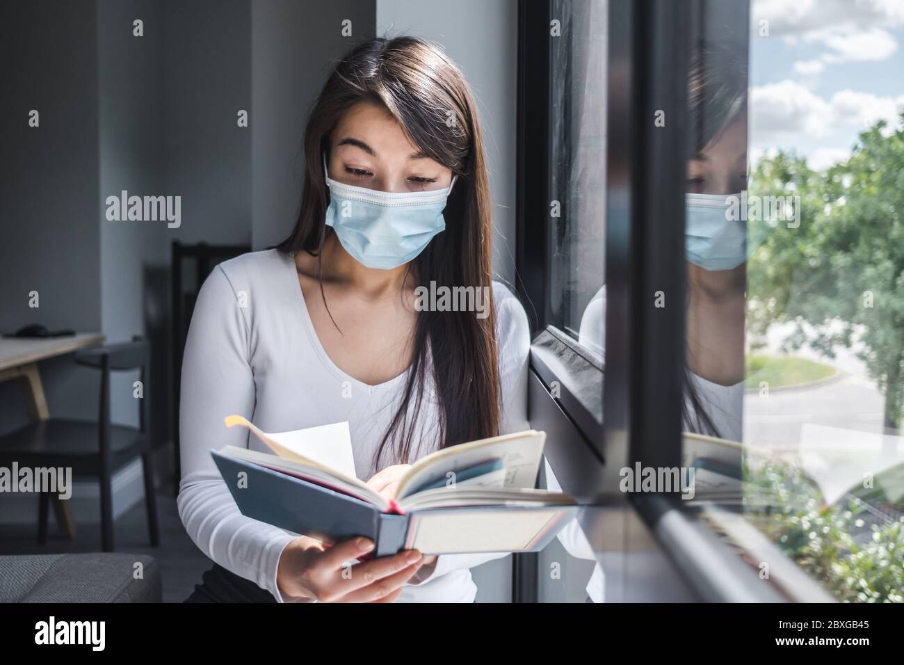 Frau mit einer Gesichtsmaske, die beim Sperren am Fenster sitzt und liest Stockfoto