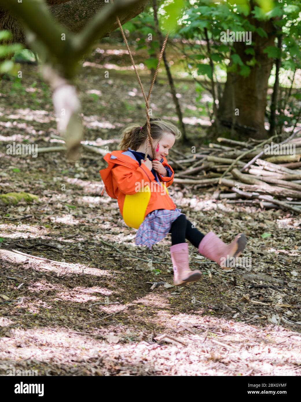 Kleines Mädchen in orangefarbenem Regenmantel, das im Wald auf Schaukel, in der Höhle, in der Nähe des Baumes spielt Stockfoto