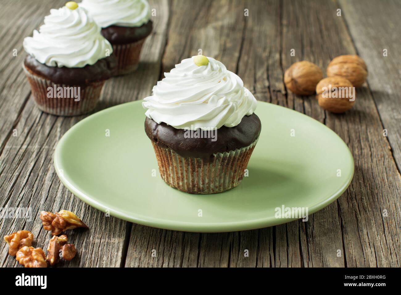 Schokoladenkuchen mit Schlagsahne. Vor dem Teller liegen Walnusskerne, zwei Cupcakes und Walnussschalen in verschwommenem Hintergrund. Stockfoto