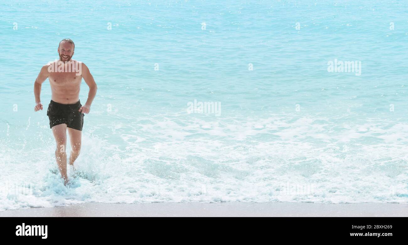 Glücklich lächelnder Mann mit perfekten Körper läuft aus Wellen Wasser spritzen. Urlaub am Meer Konzept Stockfoto