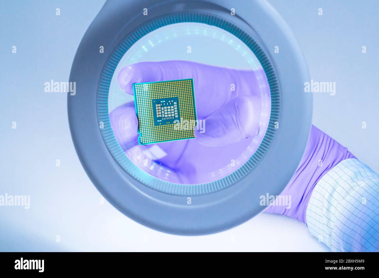 Blick auf die erste Person. Eine Hand in einem blauen Handschuh hält einen Prozessor-Chip und untersucht ihn unter einer Lupe. Stockfoto