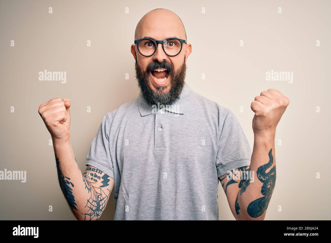 Schöner Glatze mit Bart und Tattoo in lässigem Polo und Brille, die  überrascht und erstaunt über den Erfolg mit erhobenen Armen und offenen  Augen feiert. W Stockfotografie - Alamy