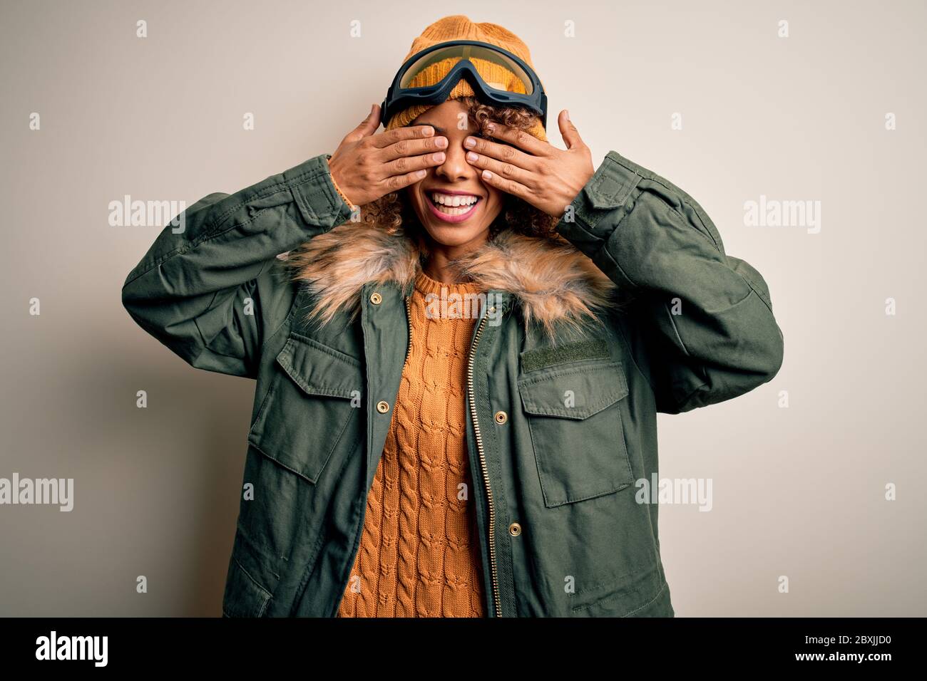 Junge afroamerikanische Skifahrer Frau mit lockigen Haaren tragen Schnee Sportbekleidung und Skibrille bedeckt Augen mit Händen lächelnd fröhlich und lustig. Blind Stockfoto
