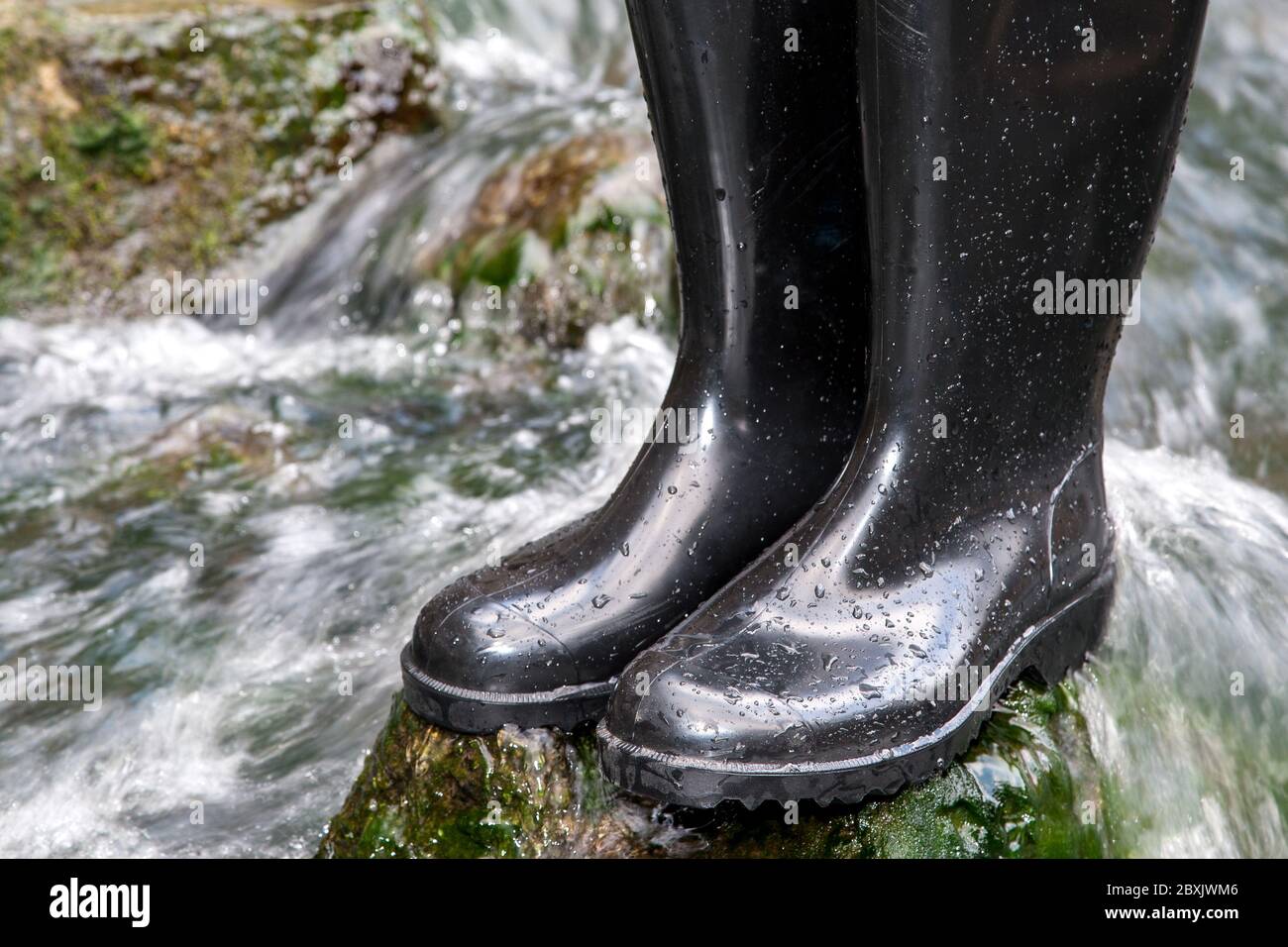 Schwarze Gummistiefel stehen auf einem moosbedeckten Stein inmitten des rauschenden Wassers eines Baches. Stockfoto