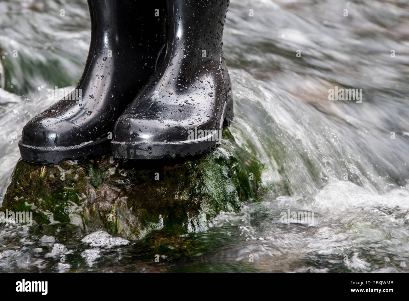 Zwei nassbesprühte Gummistiefel stehen auf einem Stein im rauschenden Wasser eines Baches. Gummistiefel halten deine Füße auch in nassem Gelände trocken. Stockfoto