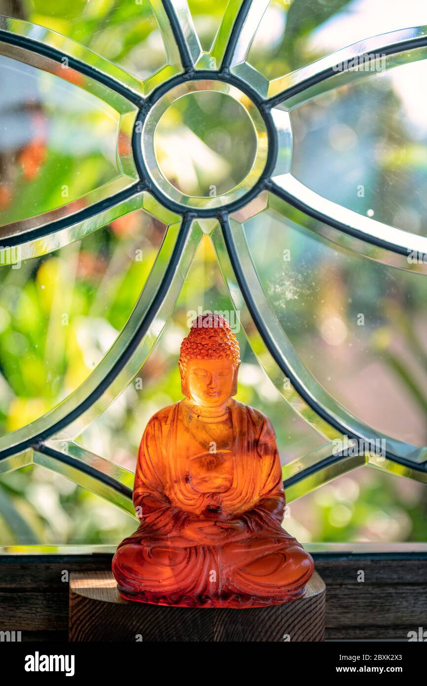 Sonnenlicht scheint durch einen Glas-Buddha, der vor einem Fenster mit Pflanzen im Hintergrund sitzt. Stockfoto