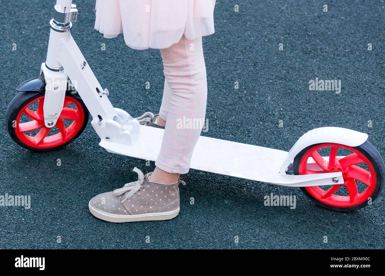 Ein kleines stilvolles Mädchen, das neben ihrem weißen Roller auf der Straße steht. Nahaufnahme. Nur Beine. Stockfoto