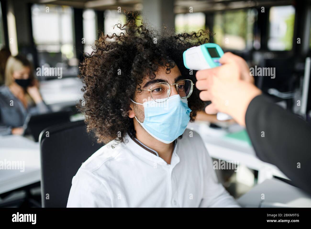 Junger Mann mit Gesichtsmaske zurück bei der Arbeit im Büro nach Lockdown, Messung der Temperatur. Stockfoto