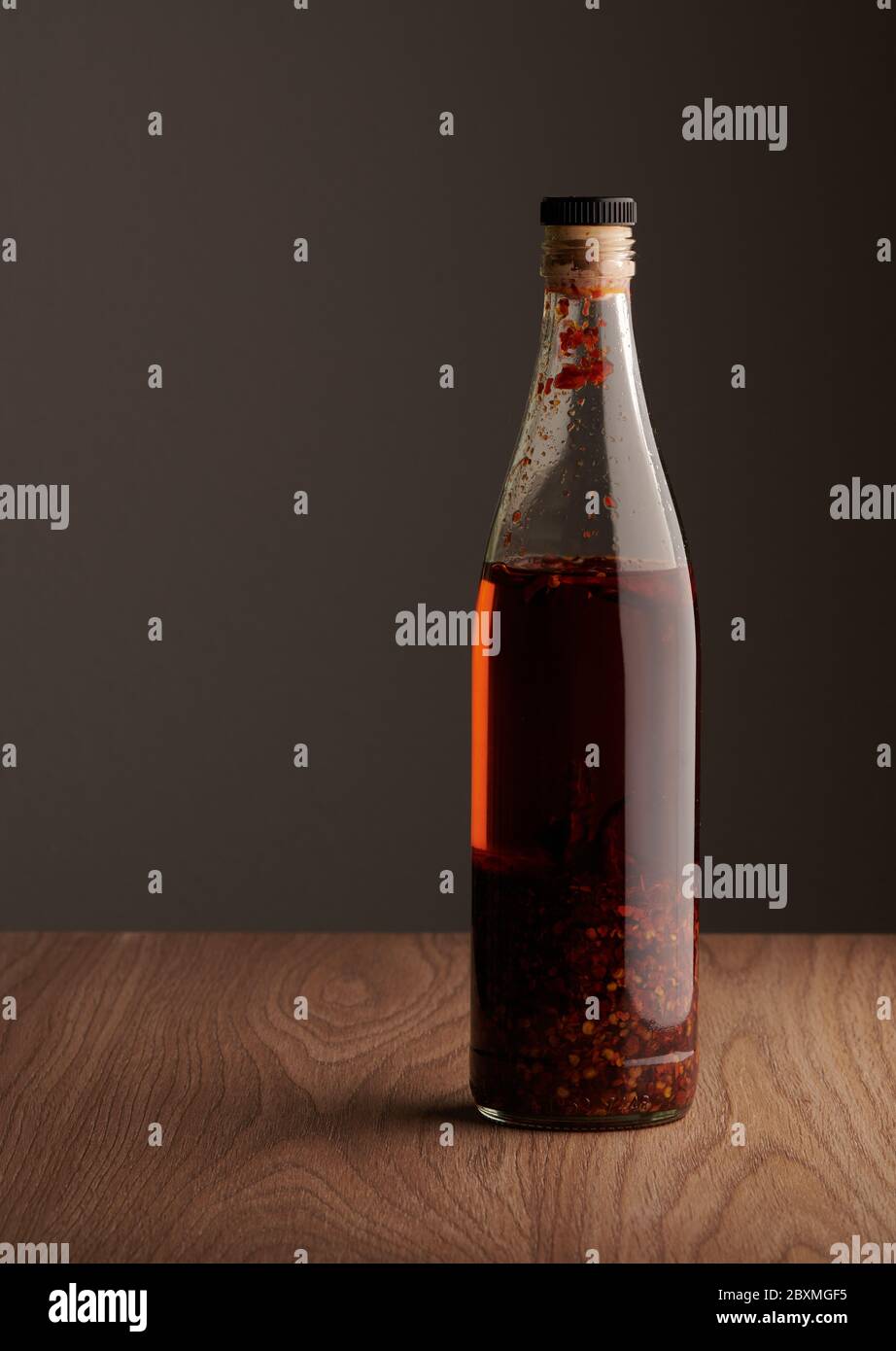 Transparente Flasche hausgemachtes Chilliöl mit Stücken von trockenem Chili, auf einem Holztisch Stockfoto