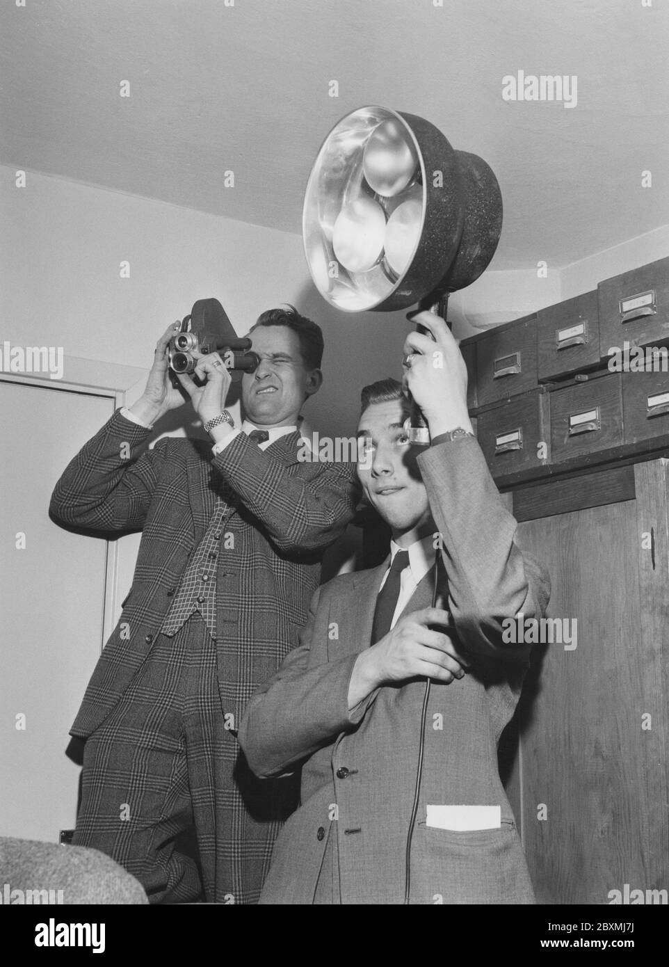 Dreharbeiten in den 1950er Jahren. Ein Mann filmt mit einer kleinen 8-mm-Filmkamera, während sein Freund Lampen hält, um das gefilmte Motiv zu beleuchten. Die Kamera produzierte einen analogen Film, der von einem Projektor auf einer weißen Leinwand angesehen wurde. Schweden 1958 Stockfoto