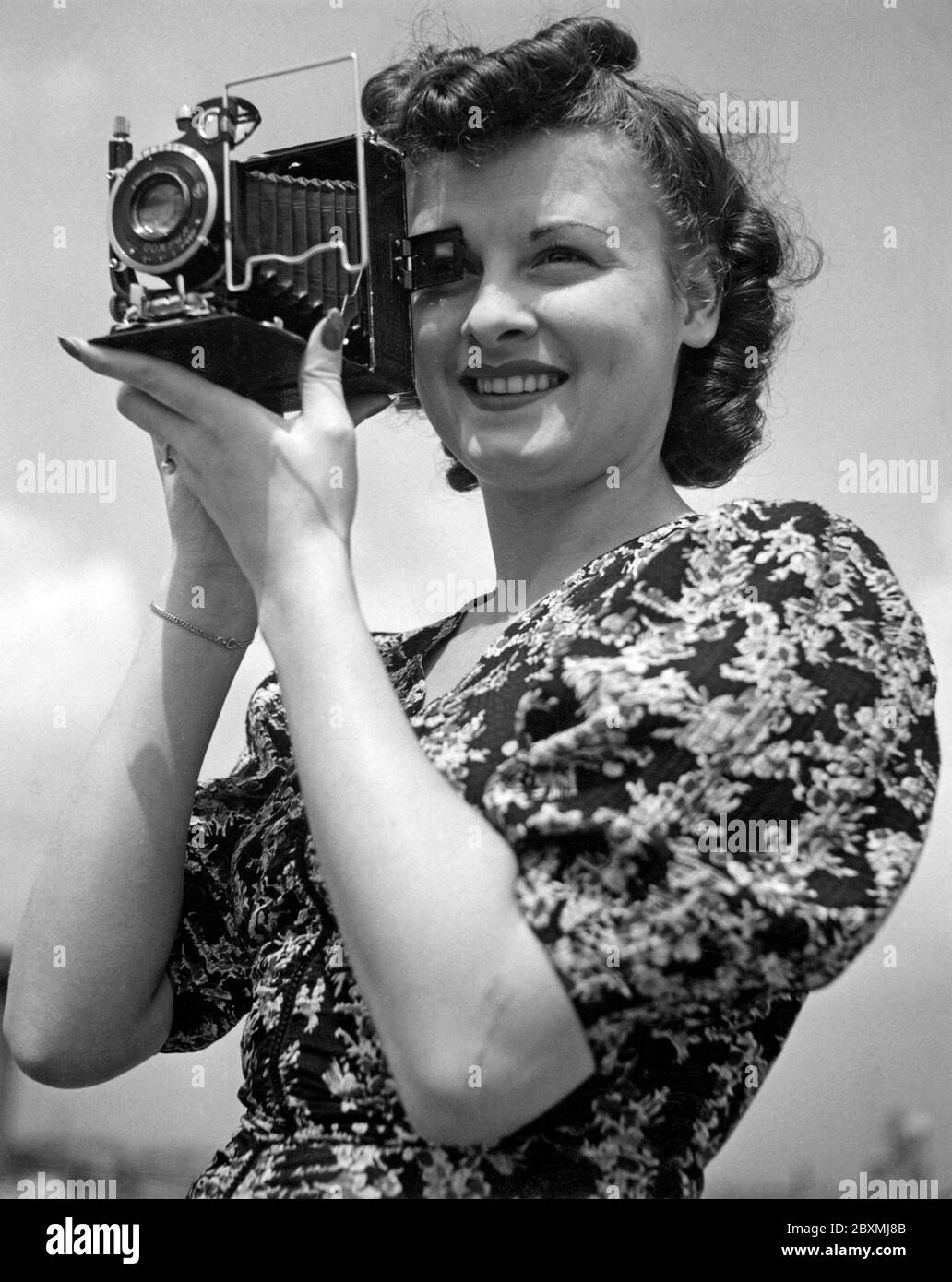 Amateurfotograf in den 1940er Jahren. Eine junge Frau fotografiert an einem Sommertag. Das Kameramodell ist praktisch. Wenn Sie es nicht verwenden, werden die Linse und der Balg in das Kameragehäuse gefaltet. Um Bilder aufzunehmen, haben Sie es geöffnet und waren bereit zu gehen. Die Kamera produzierte analogen Film. Stockfoto