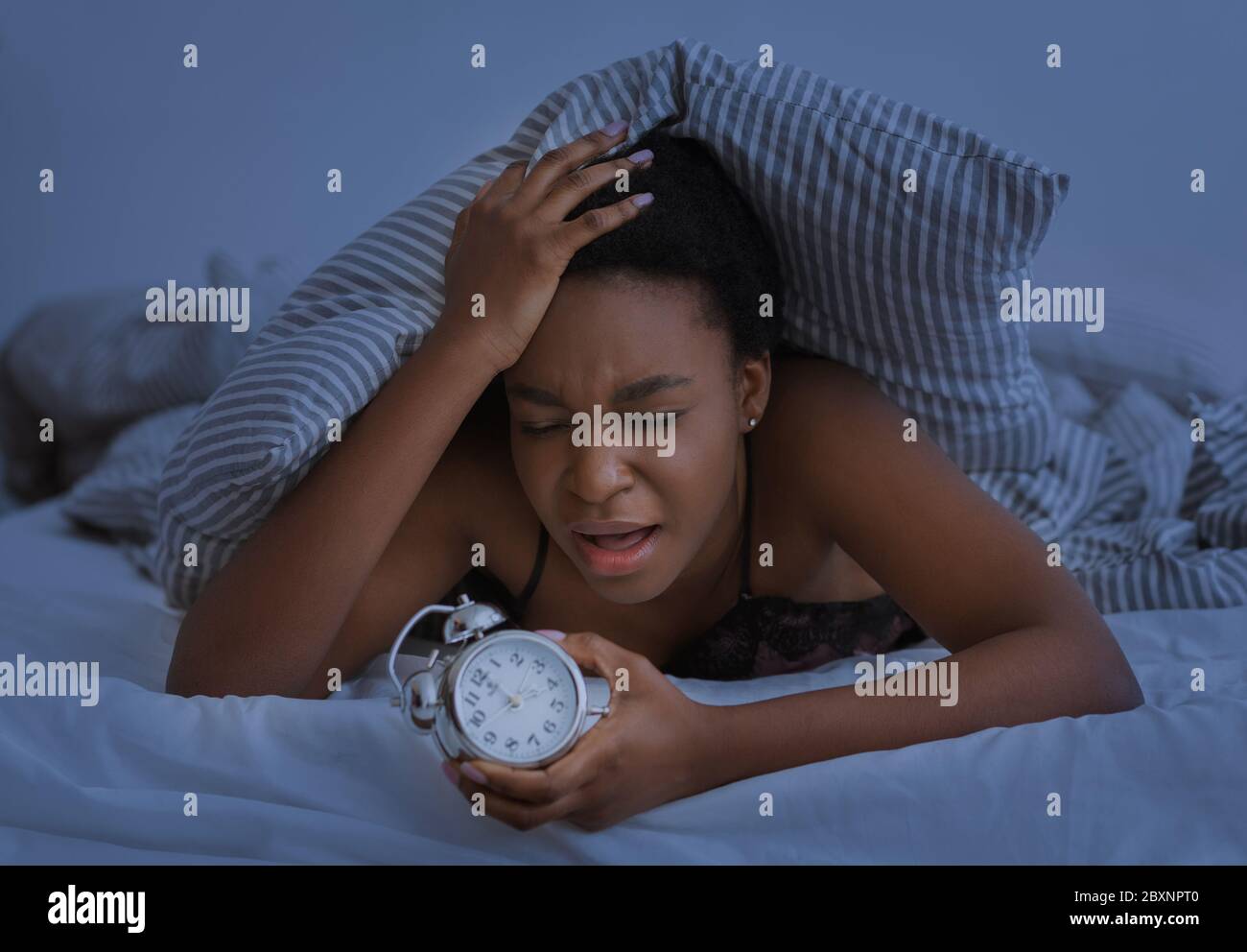 Schlaflose Nacht. Traurig afroamerikanischen Mädchen drückt Hand an Kopf und hält Wecker Stockfoto
