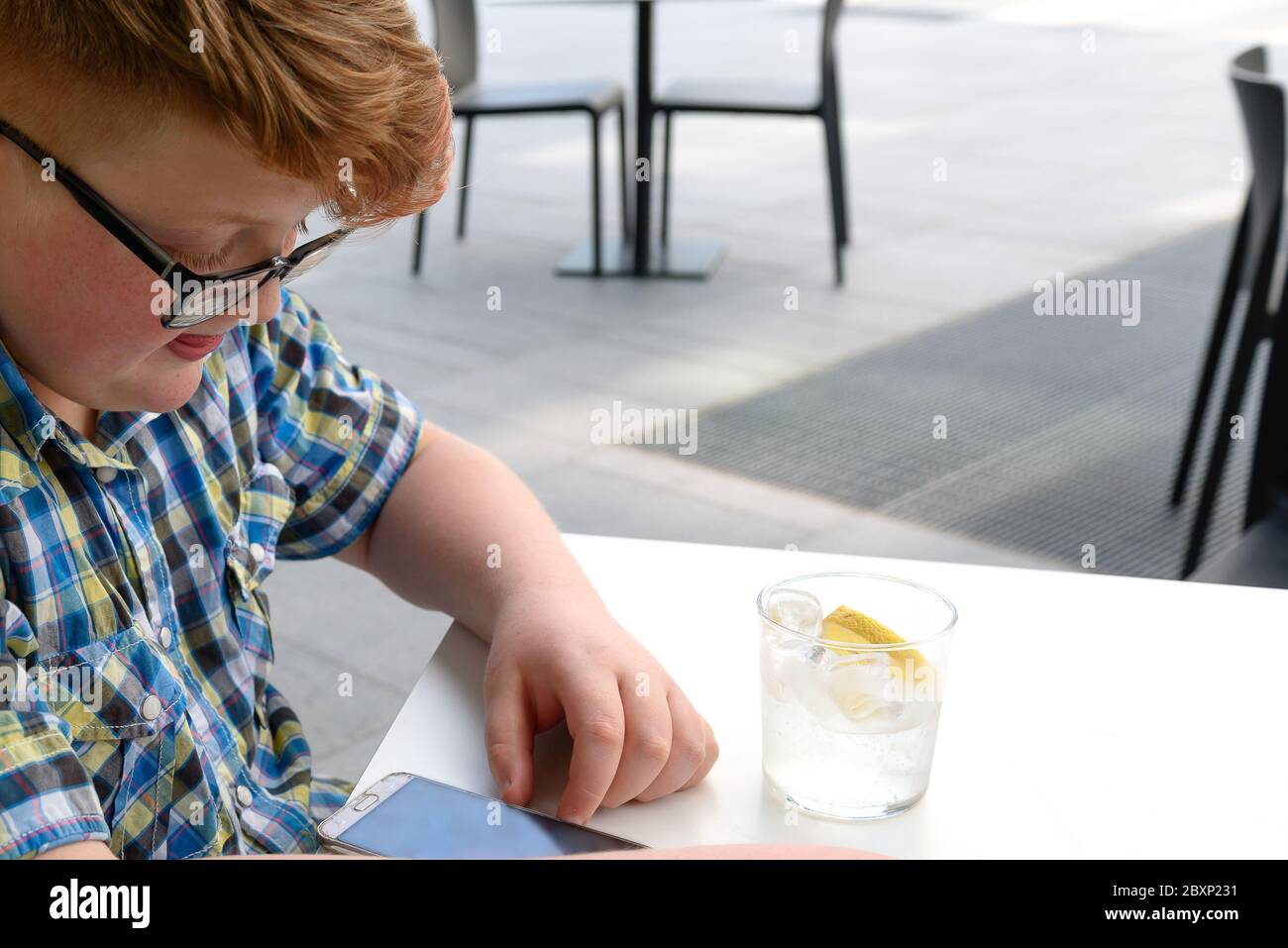 Rothaariger Junge mit Handy, der aus einem Glas Eiswasser mit Zitrone trinkt. Kind mit Brille in einem karierten Hemd gekleidet sieht auf das Smartphone und d Stockfoto