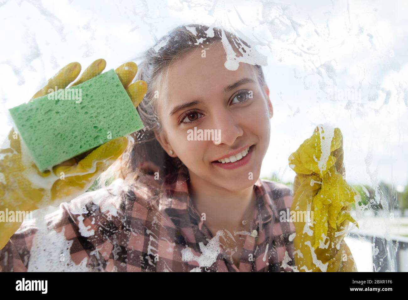 Reinigungshaus Concepta junges Mädchen mit einem Lächeln in gelben Gummihandschuhen wäscht ein Fenster von außen. Stockfoto